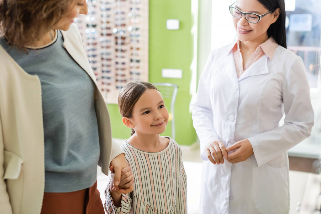 pleased mother and daughter holding hands near asian oculist in optics store - Photo, Image