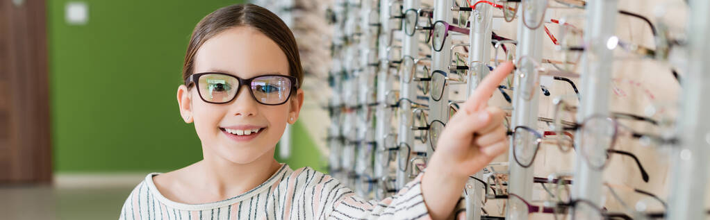 joyful girl looking at camera while choosing eyeglasses in optics store, banner - Foto, Bild