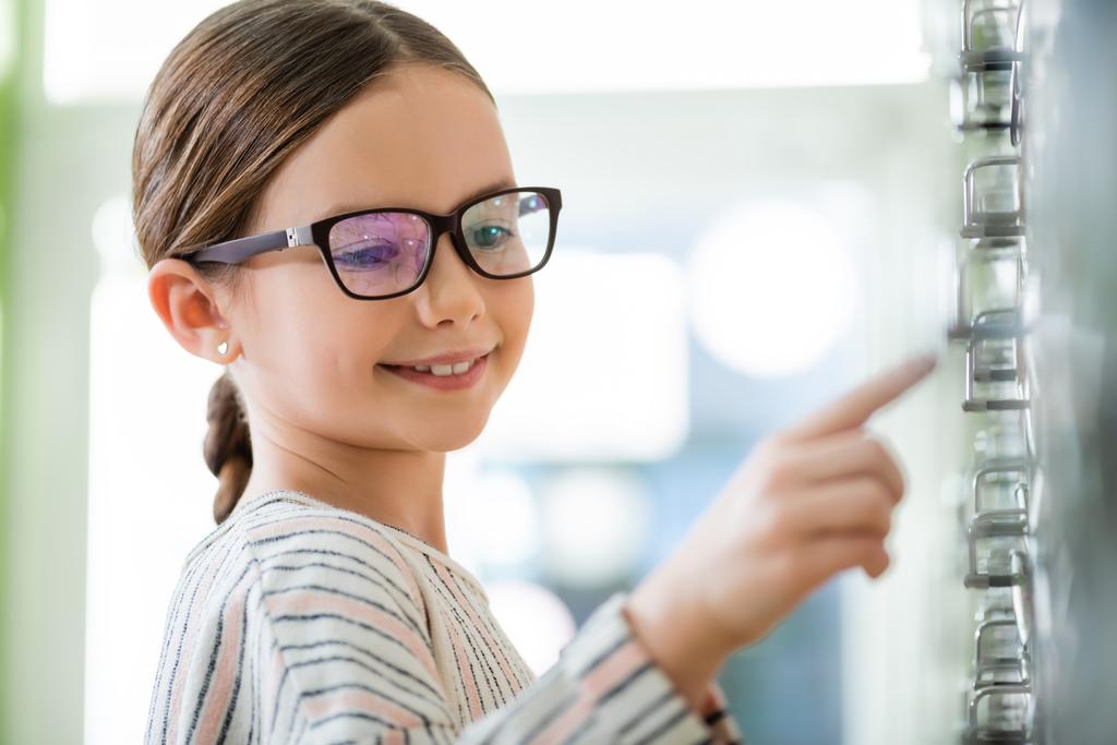 smiling girl in eyeglasses pointing with finger in optics store on blurred foreground - Photo, image