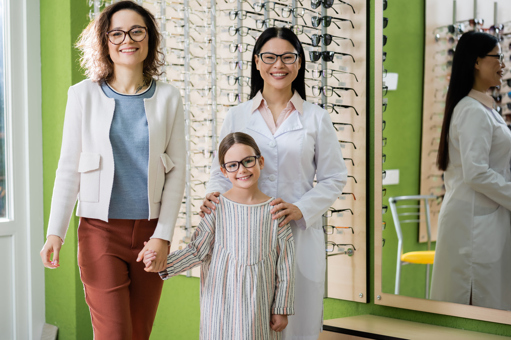 happy mother, daughter and asian oculist in eyeglasses looking at camera in optics salon - Fotoğraf, Görsel