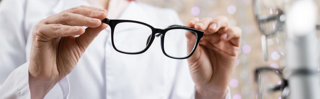 обрізаний вид окуліста в білому пальто, що тримає окуляри в салоні оптики, банер
 - Фото, зображення
