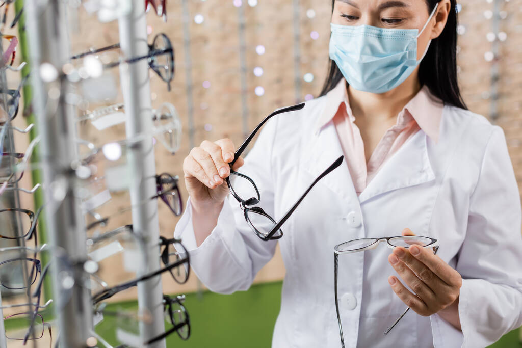 oftalmólogo asiático en máscara médica comparando gafas en tienda de óptica - Foto, Imagen