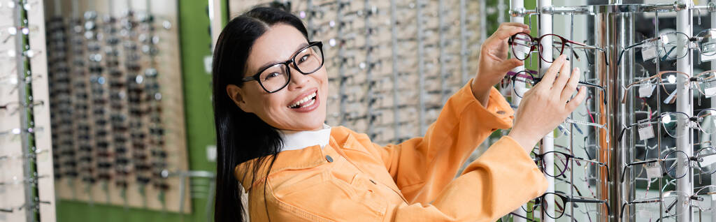excited asian woman choosing eyeglasses and looking at camera in optics store, banner - Foto, Bild