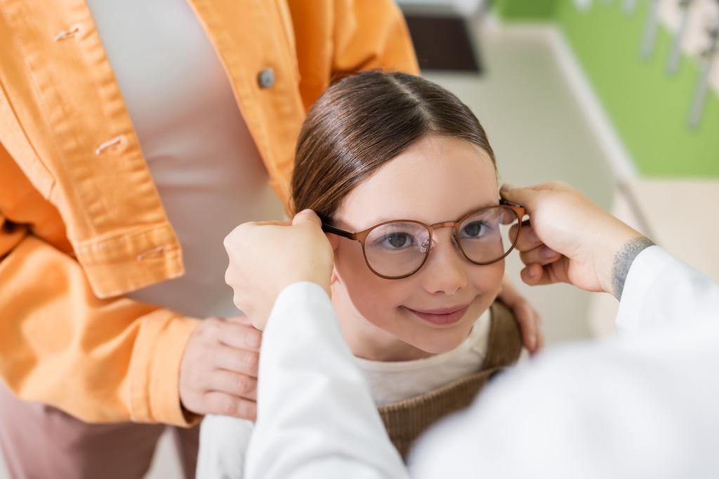 blurred ophthalmologist putting glasses on positive girl in optics store - Foto, immagini