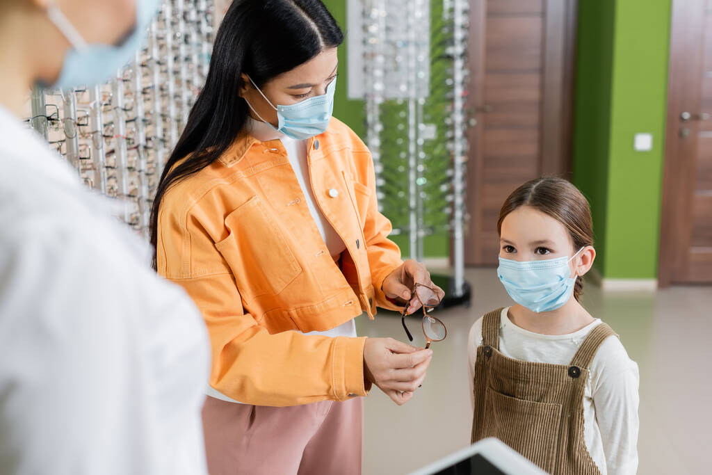 asian woman in medical mask holding eyeglasses near daughter and blurred oculist in optics store - Zdjęcie, obraz