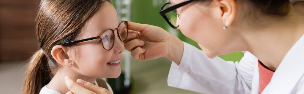 οφθαλμίατρος δοκιμάζει γυαλιά σε χαμογελαστό κορίτσι στο κατάστημα οπτικών, πανό - Φωτογραφία, εικόνα