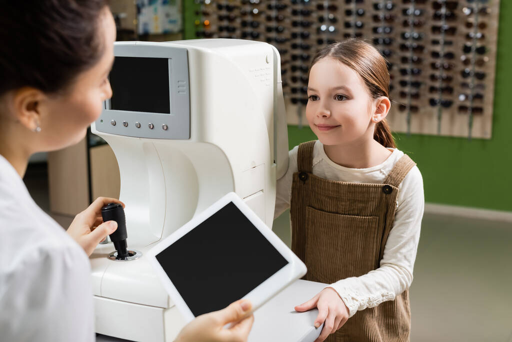 blurred oculist holding digital tablet with blank screen near positive girl and vision screener in optics store - Foto, afbeelding
