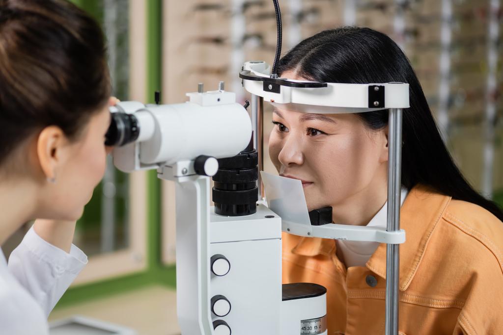 blurred optometrist testing eyesight of asian woman on vision screener in optics shop - Foto, immagini