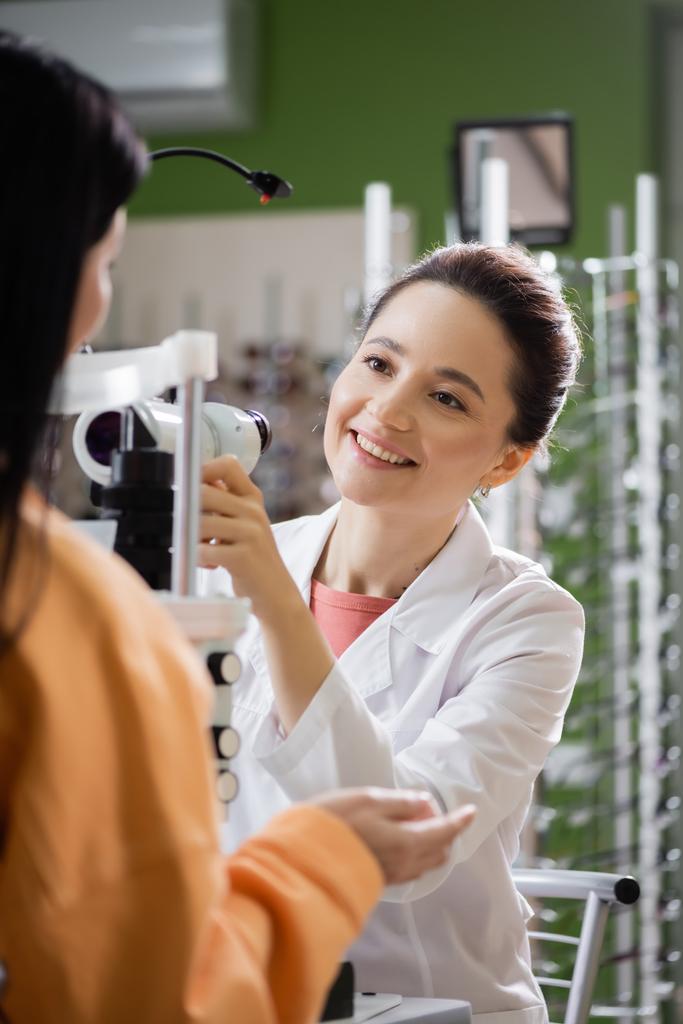 νέος οπτομέτρης χαμογελώντας κοντά σε θολή γυναίκα και οφθαλμοσκόπιο στο κατάστημα οπτικών - Φωτογραφία, εικόνα
