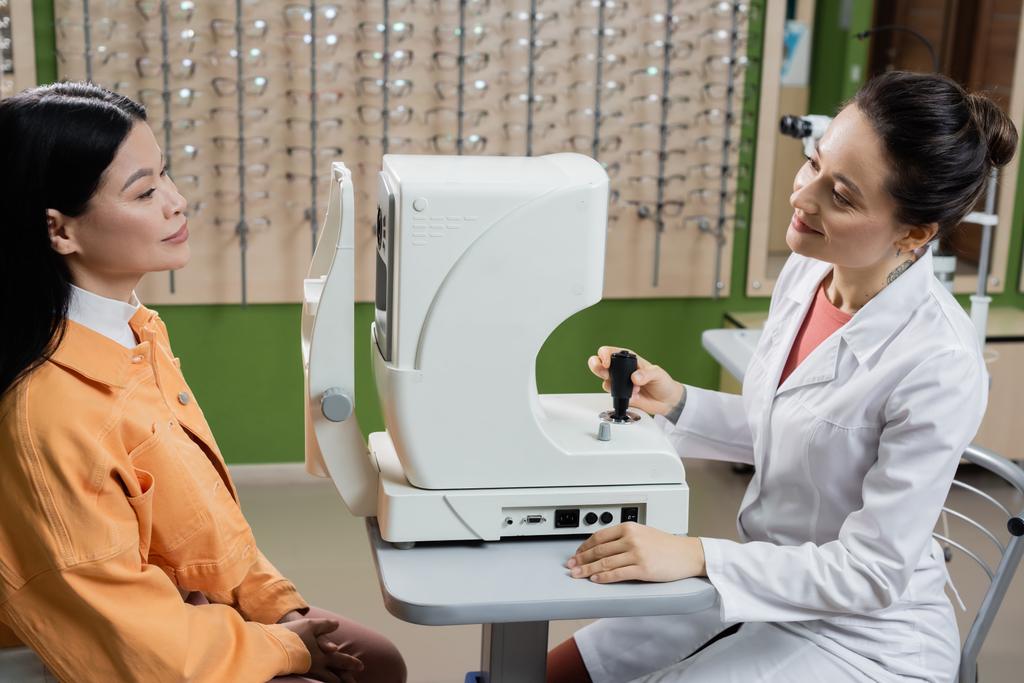 sonriente optometrista mirando asiático mujer cerca visión cribador y borrosa surtido de gafas - Foto, Imagen