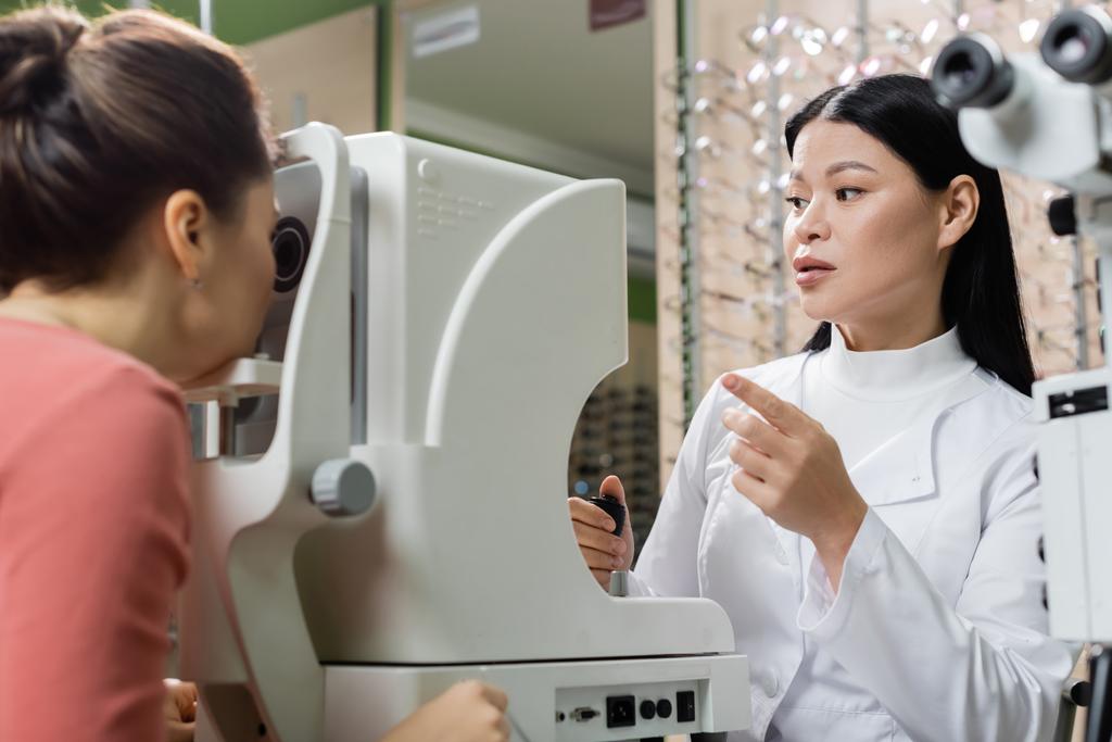 asian optometrist pointing with finger while measuring eyesight of blurred woman on vision screener - Foto, imagen