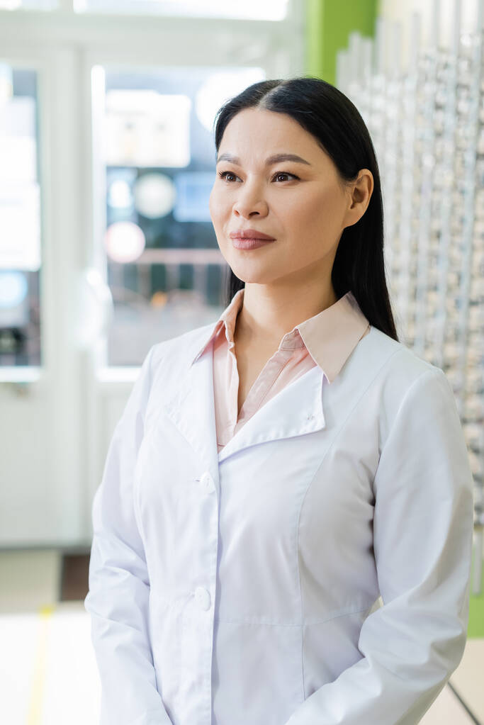 ophtalmologiste asiatique en manteau blanc debout dans la boutique d'optique sur fond flou - Photo, image
