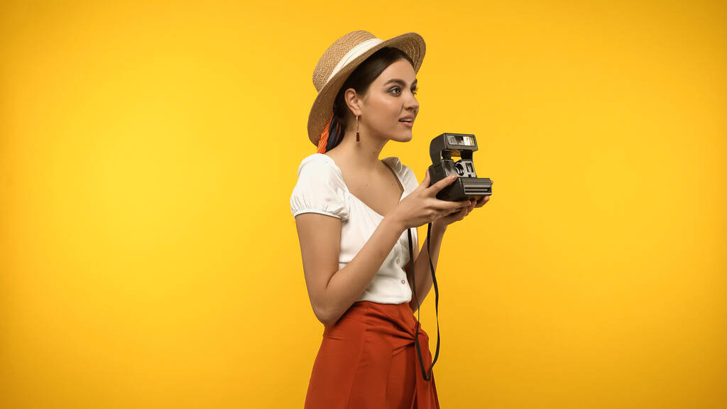 Χαμογελαστή γυναίκα με ψάθινο καπέλο κρατώντας vintage κάμερα απομονωμένη στο κίτρινο - Φωτογραφία, εικόνα