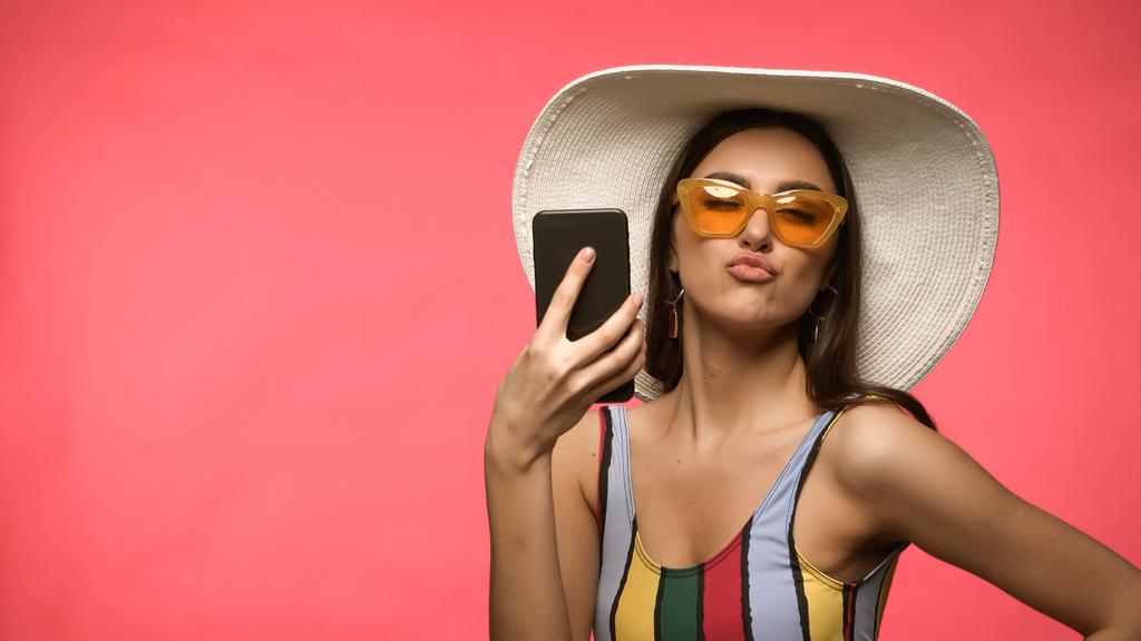 Mladá žena v plavkách a slunečních brýlích drží mobilní telefon a našpulené rty izolované na růžové  - Fotografie, Obrázek
