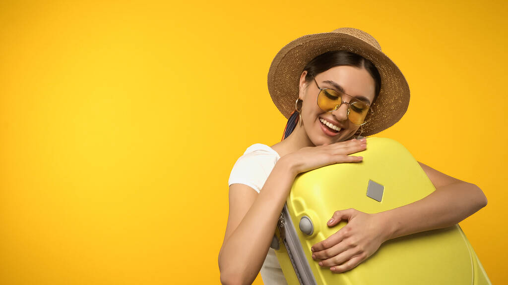 Young tourist in sun hat and sunglasses holding suitcase isolated on yellow  - Foto, Imagen