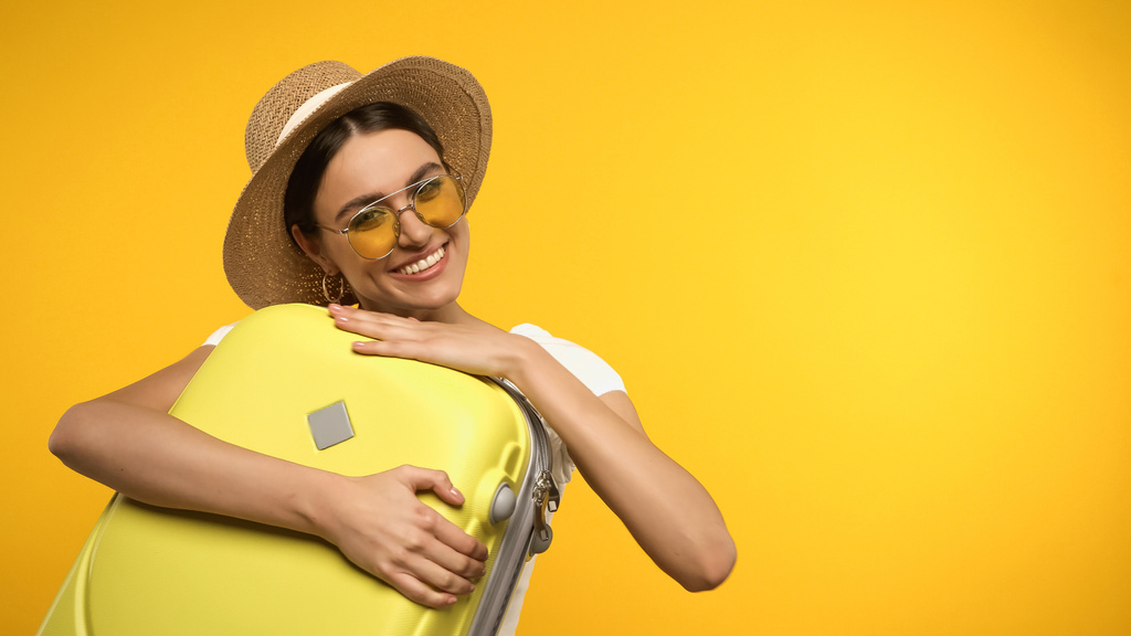 Young woman in sunglasses hugging suitcase isolated on yellow  - Photo, Image