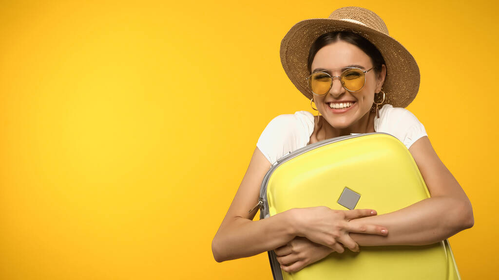 Reisender mit Strohhut umarmt Koffer isoliert auf gelb  - Foto, Bild