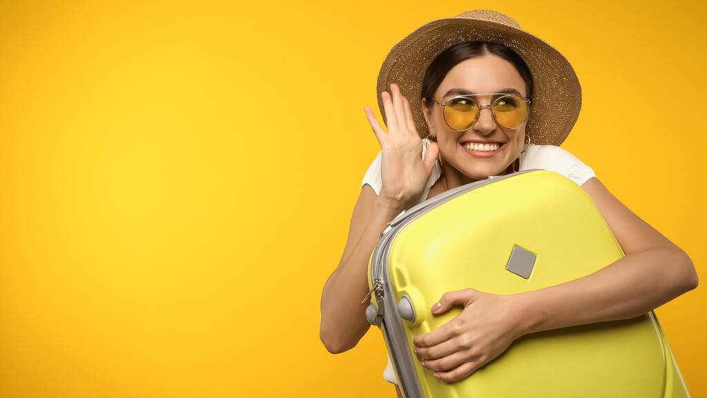 Turista sonriente en gafas de sol sosteniendo la maleta y agitando la mano aislado en amarillo  - Foto, imagen