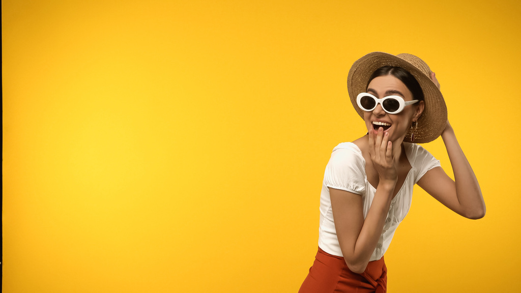 Excited brunette woman in straw hat and sunglasses isolated on yellow  - 写真・画像