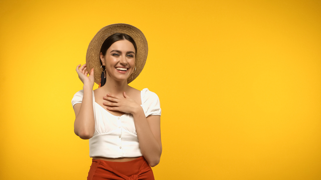 Smiling model in blouse and straw hat looking at camera isolated on yellow  - Фото, изображение