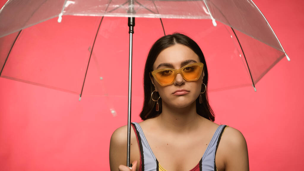 Sad woman in swimsuit and sunglasses holding umbrella isolated on pink  - Foto, Imagem