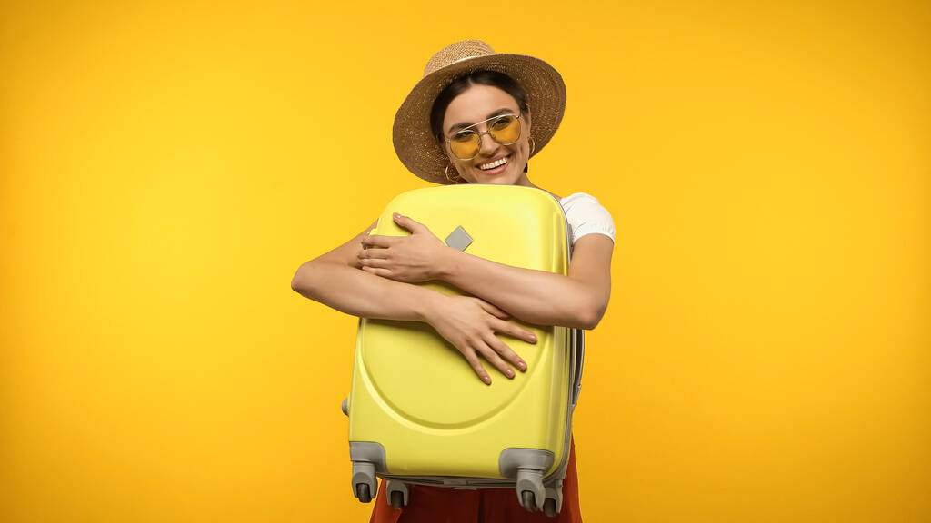 Joven viajero en gafas de sol abrazando maleta aislada en amarillo  - Foto, imagen
