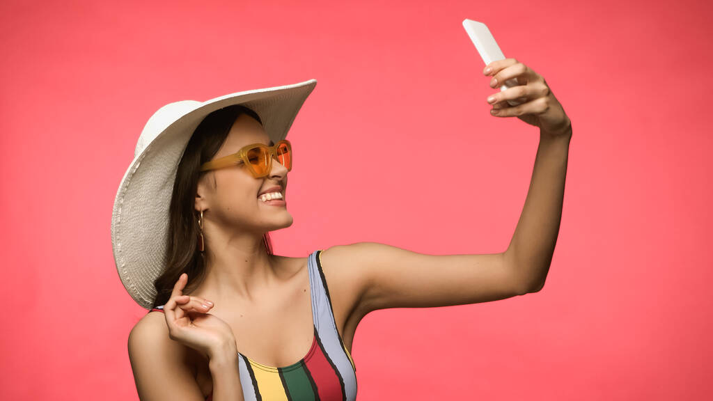 Onnellinen nainen aurinkohattu ottaen selfie älypuhelimeen eristetty vaaleanpunainen  - Valokuva, kuva