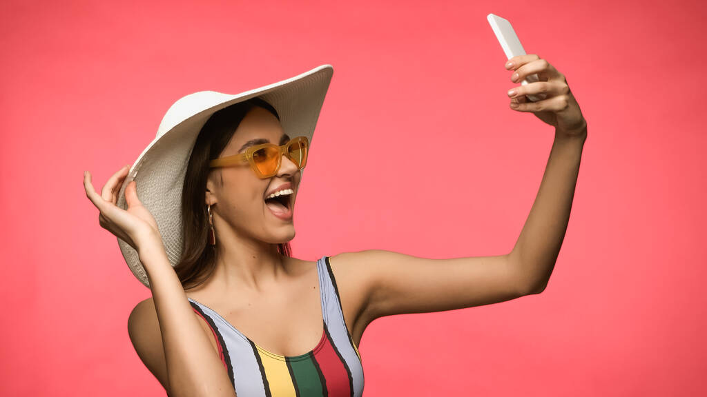 Ενθουσιασμένη γυναίκα με γυαλιά ηλίου και μαγιό που παίρνει selfie σε smartphone που απομονώνεται σε ροζ  - Φωτογραφία, εικόνα