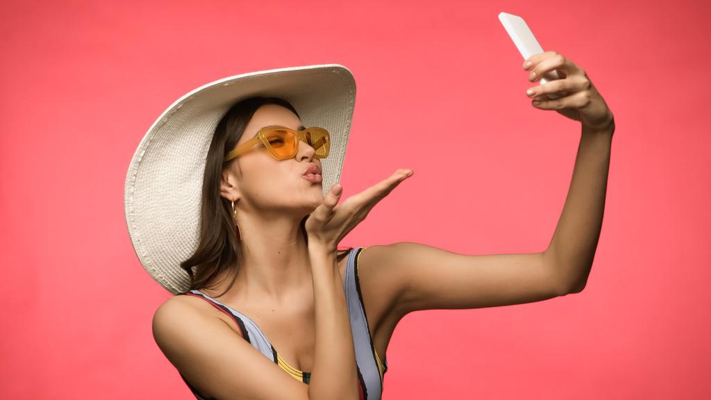 Young model in swimsuit taking selfie on smartphone and blowing air kiss isolated on pink  - Photo, Image