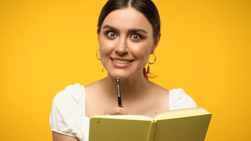 Brunette woman looking at camera while holding pen near notebook isolated on yellow - Фото, изображение