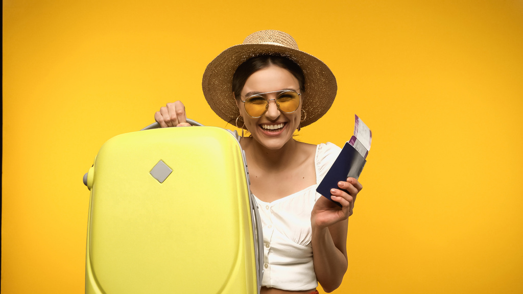 Χαμογελαστή τουρίστας σε ψάθινο καπέλο παραδιδόμενες αποσκευές και διαβατήριο που απομονώνονται σε κίτρινο - Φωτογραφία, εικόνα