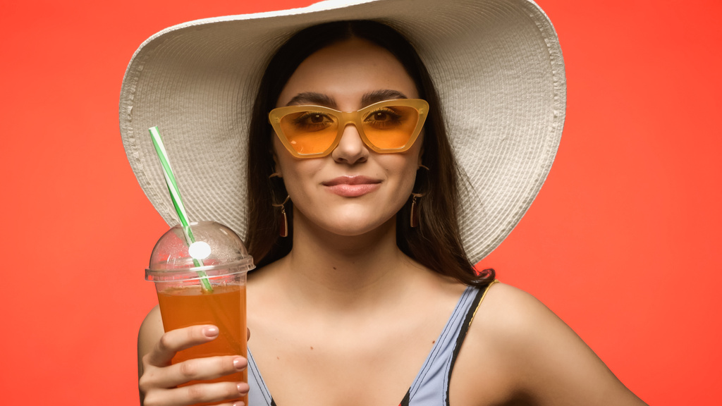 Pretty brunette woman in sun hat holding cocktail in plastic cup isolated on coral  - 写真・画像