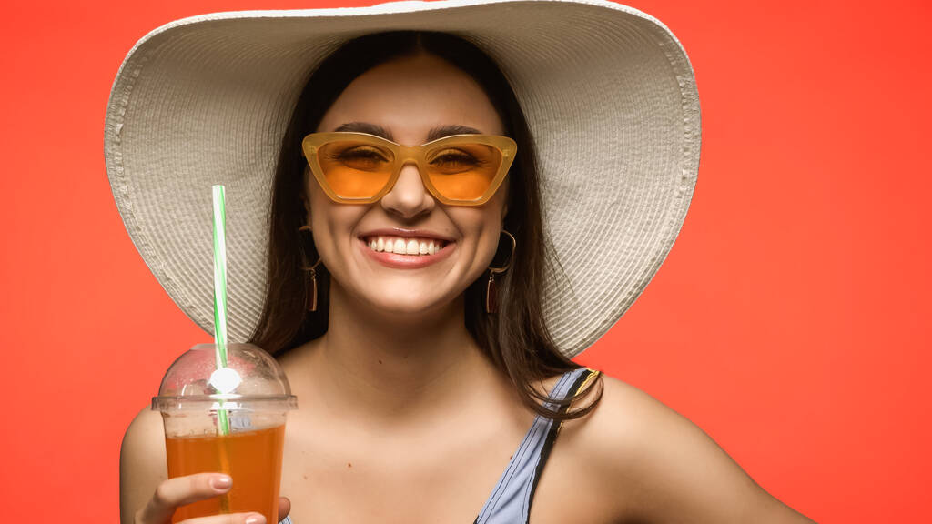 Pretty model in straw hat holding cocktail in plastic cup isolated on coral  - Fotografie, Obrázek