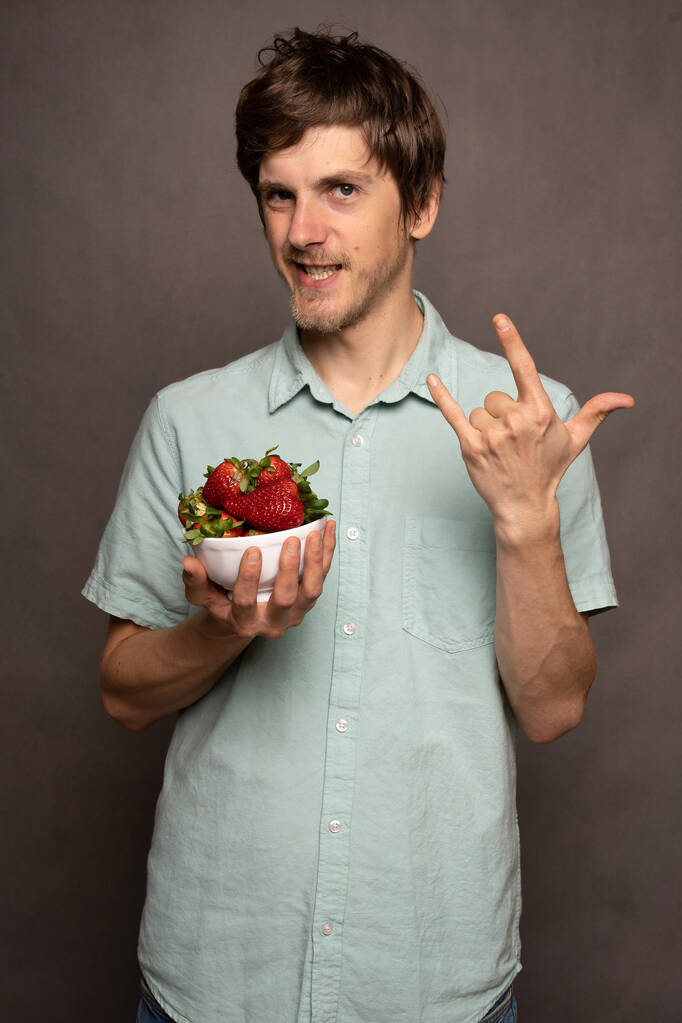 Νέος όμορφος ψηλός λεπτός λευκός άνδρας με καστανά μαλλιά κρατώντας φράουλες κάνοντας τα κέρατα σημάδι σε γαλάζιο πουκάμισο σε γκρι φόντο - Φωτογραφία, εικόνα