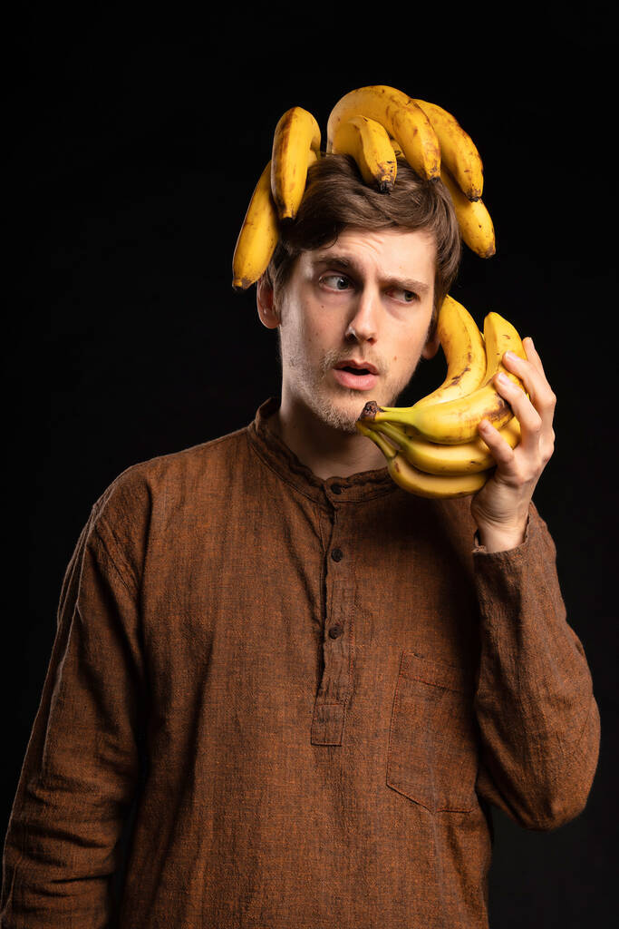 Young handsome tall slim white man with brown hair using bananas as phone with brown shirt on black background - Valokuva, kuva