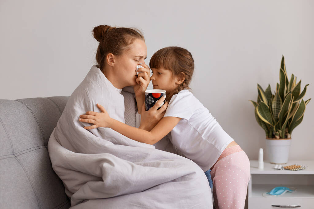 Profile portrait of sick unhealthy woman with runny nose and high temperature drinking hot tea, charming little daughter hugging mother, wants her to feel better. - Photo, Image