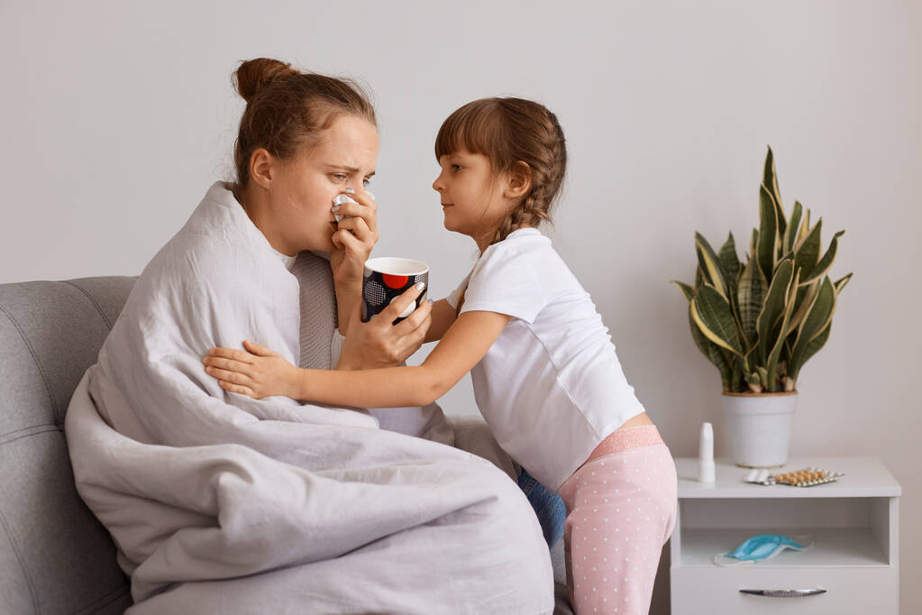 Indoor shot of unhealhty woman drinking tea and suffering runny nose, cute child hugging sick mother, taking care of her mom, posing on light room at home. - Photo, Image