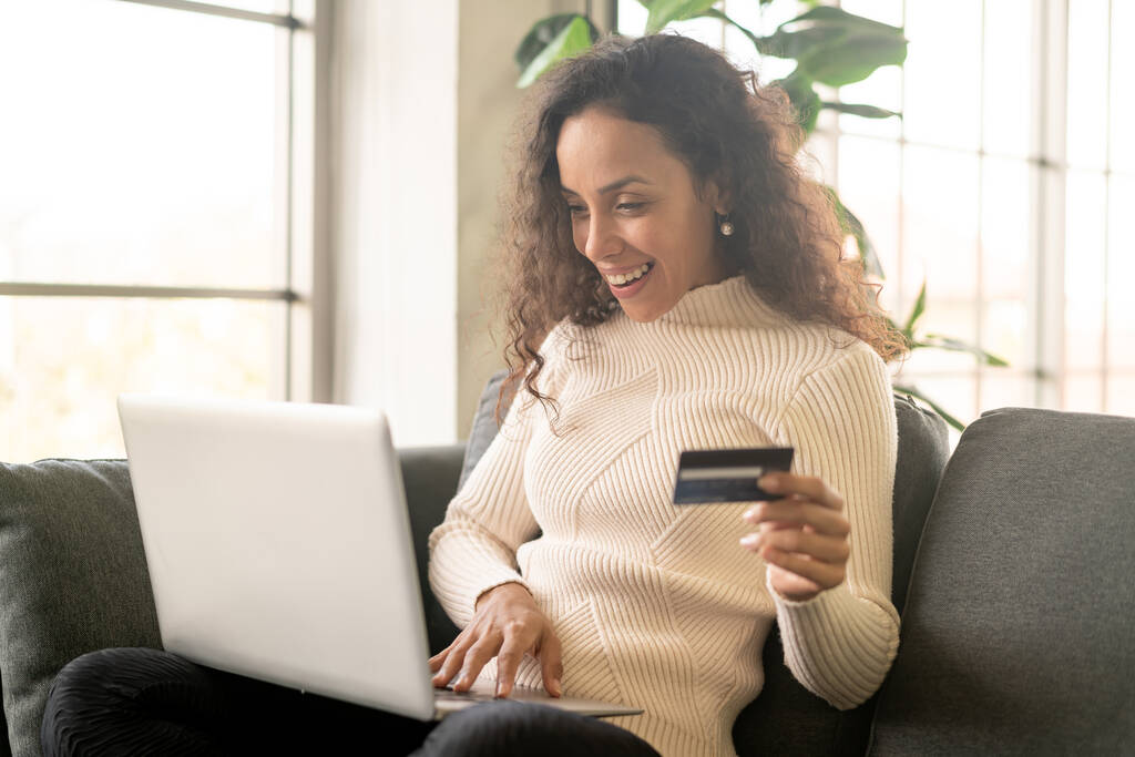 Evdeki kanepede alışveriş yapmak için dizüstü bilgisayar ve kredi kartı kullanan Latin bir kadın. - Fotoğraf, Görsel