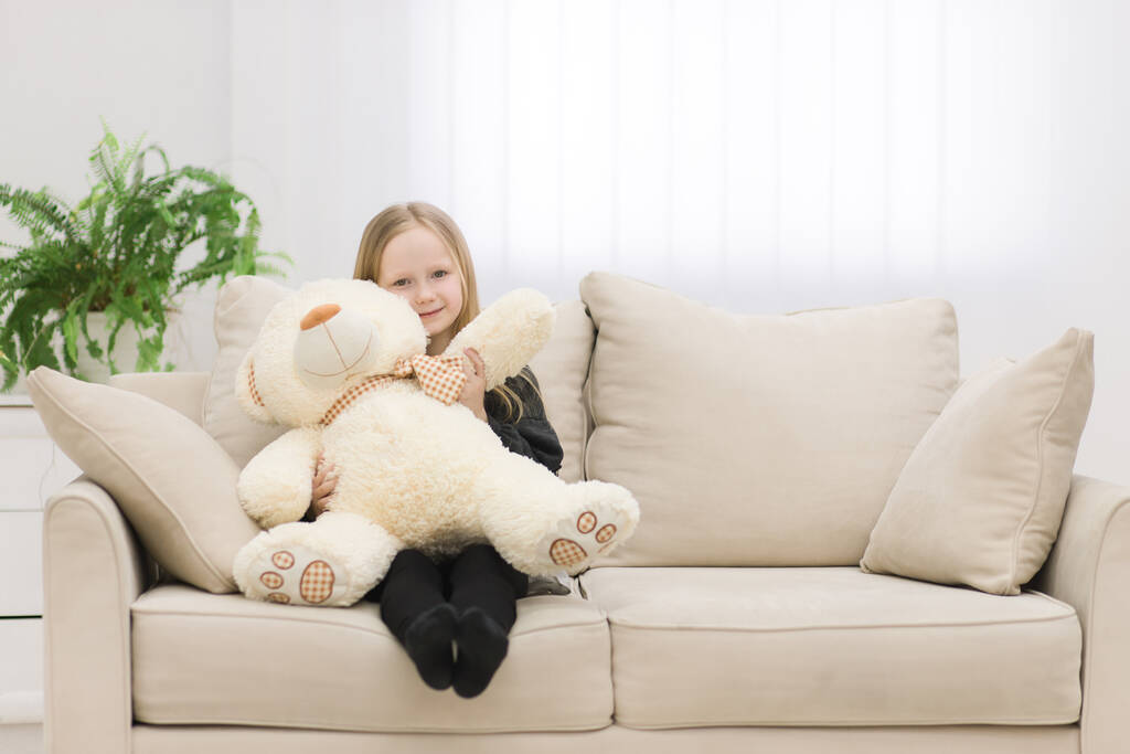 Photo of cute little girl sitting with teddy bear on white sofa. - Zdjęcie, obraz