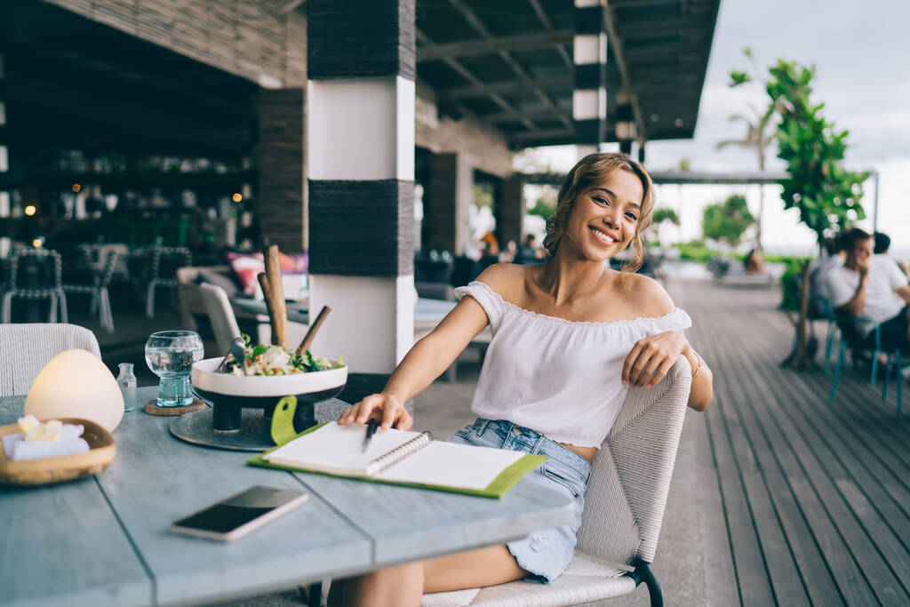 Χαμογελαστή γυναίκα ελεύθερη επαγγελματίας φορώντας καλοκαιρινά ρούχα κάθεται σε ξύλινο τραπέζι σε βεράντα εστιατόριο με πιάτο στυλό σημειωματάριο νερού, ενώ κοιτάζοντας την κάμερα - Φωτογραφία, εικόνα