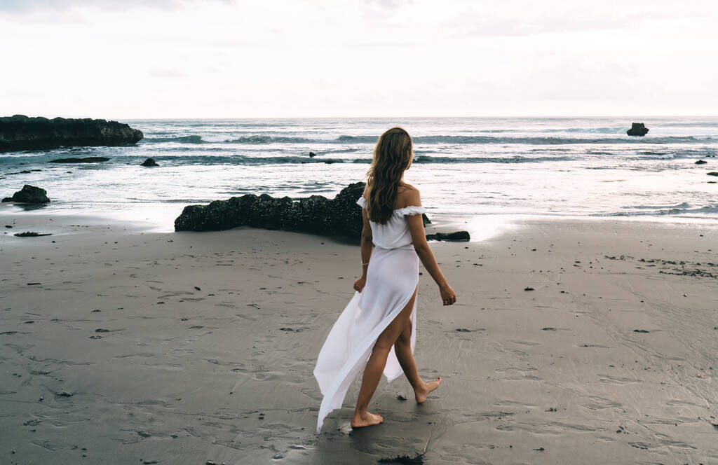 Вид ззаду анонімного молодого босоногих туристів з довгим волоссям у сукні максі, що йде на піщаному пляжі до махаючого океану і насолоджується часом заходу сонця
 - Фото, зображення