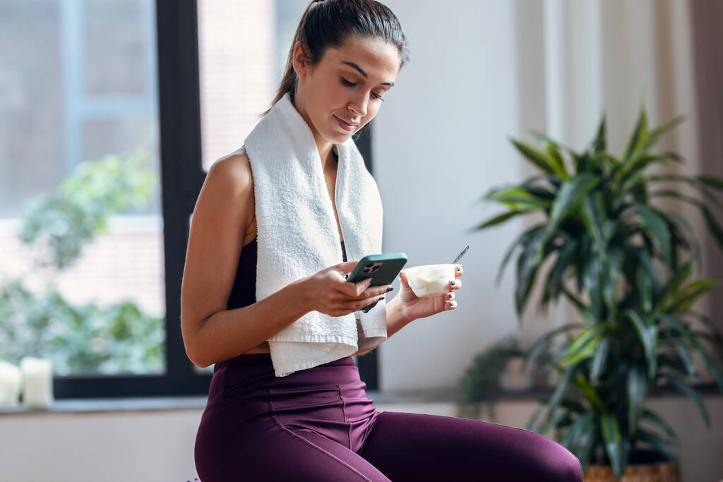 Prise de vue d'une jeune femme sportive mangeant un yaourt en utilisant un smartphone assis sur un ballon de fitness à la maison. - Photo, image
