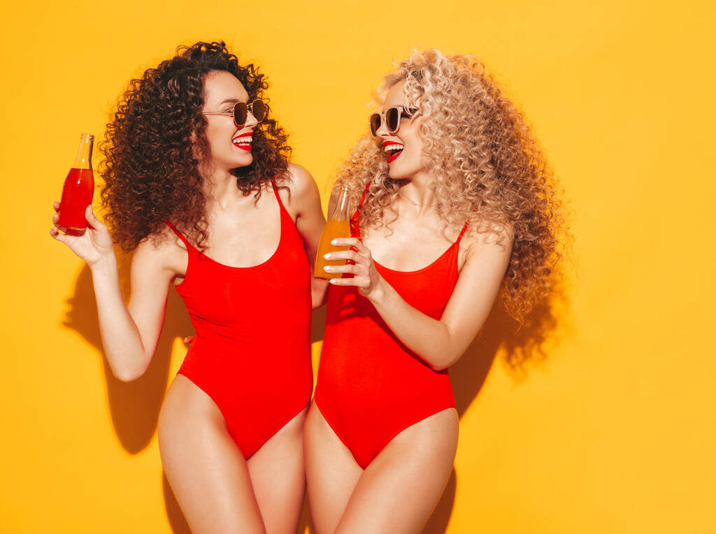 Дві красиві сексуальні усміхнені жінки хіпстера в червоному літньому купальні костюми.Трендові моделі з зачіскою з афро завитками розважаються в студії.Гаряча жінка ізольована на жовтому. Випивання лимонаду з пляшки
 - Фото, зображення