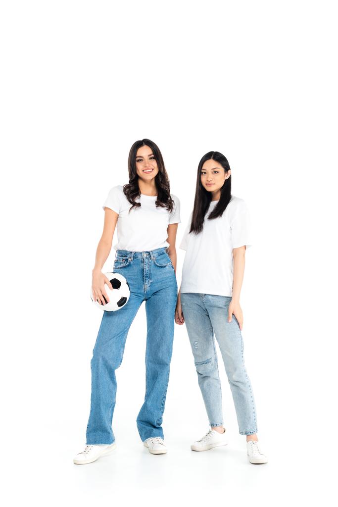 full length view of brunette woman in jeans holding ball near asian friend on white - 写真・画像