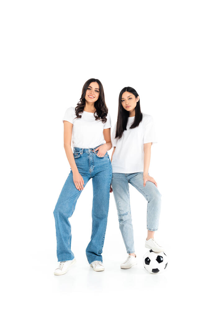 full length view of happy interracial women in jeans standing near soccer ball on white - Foto, Bild