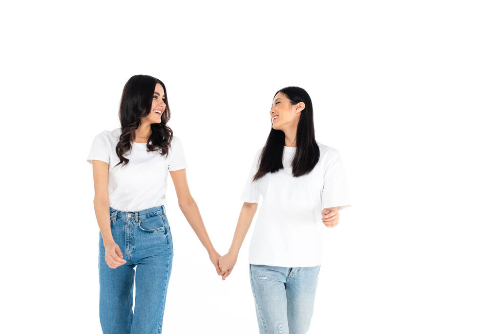 fröhliche interrassische Frauen, die sich an den Händen halten und einander isoliert auf weiß anschauen - Foto, Bild