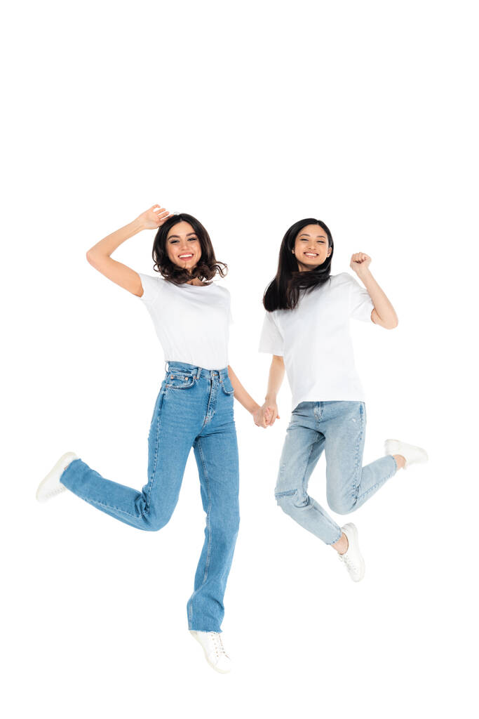 overjoyed interracial women in jeans holding hands while levitating isolated on white - Fotó, kép