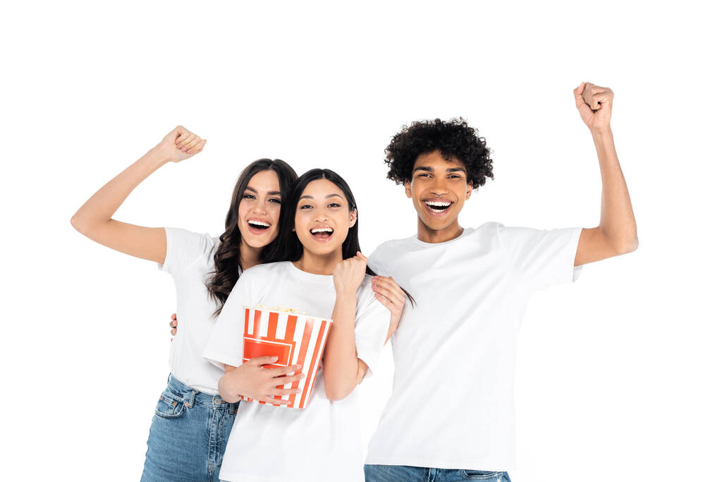 vzrušení multietničtí přátelé ukazující triumfální gesto poblíž kbelíku popcornu izolovaného na bílém - Fotografie, Obrázek