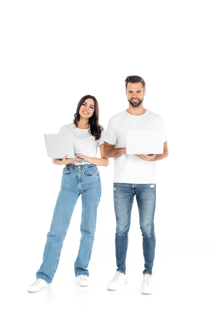 full length view of happy couple in jeans using laptops while looking at camera on white - Fotó, kép