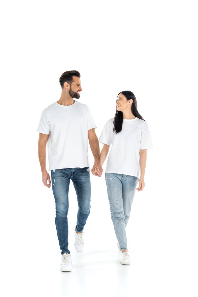 vista completa de la feliz pareja interracial mirándose el uno al otro y tomándose de la mano mientras camina sobre blanco - Foto, Imagen